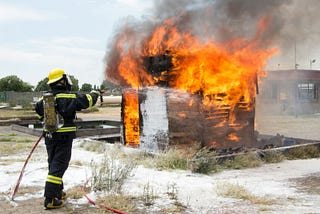 A firefighter sprays water from a hose on a small burning building.