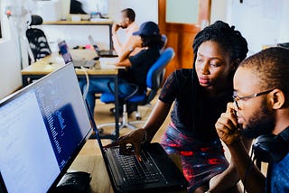 A young Black woman in a black shirt and colorful skirt pointing at a computer screen while talking to a young Black man with glasses and headphones, wearing a blue shirt. They are in an office with other employees visible in the background.