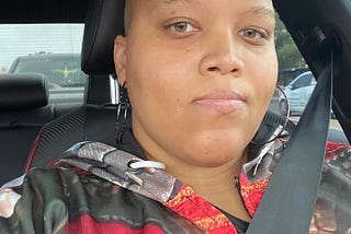 selfie of a light skinned Black woman in a car. She is looking at the camera with a blank expression. Her head is bald, and she’s wearing a hoodie that appears to have a red, brown, and gray pattern in the style of women’s armor.