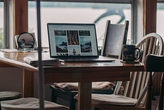 An open laptop computer sits on a kitchen table by a window overlooking a lake. A coffee cup and a book are beside it.