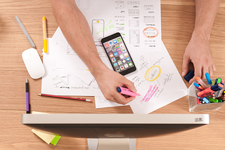 A computer and phone on a wooden desk with a person’s arm writing on papers on the desk.