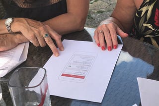 Photograph of a user research session, there is a paper prototype on the table with the hands of two women who are discussing their thoughts around the prototype.