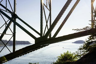 Steel bridge supports intersecting above calm water, framing open space beyond. A load-bearing structure holding tension while enabling movement beneath.