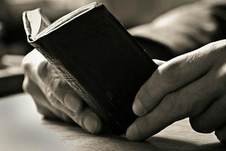 A very small, antique, hardback book is held open on a desk by the reader’s hands.