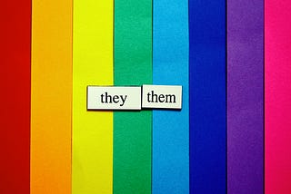 The words “they” and “them” appear over construction paper lined up to show the colors of a rainbow.