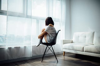woman sitting on plastic chair in living room, looking out a large window, hugging her knees