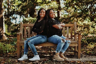 Young adult twins wearing jeans and black shirts sit with their backs together on a wooden bench. Trees are in the background.