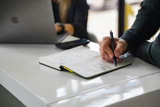 The image shows of two colleagues working together on a table in an office. The male employee is writing some notes about the discussion that they are having at the moment