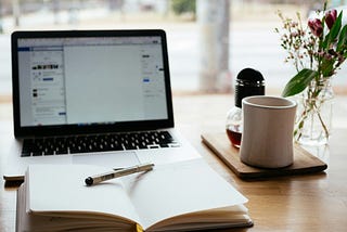laptop with an open journal in front of it and a coffee mug to the right.