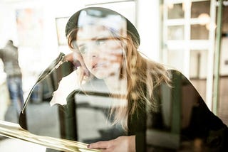 A young emerging adult wearing a black leather jacket and black hat leaning on her right arm looking out the window thinking — consulting with her future self.