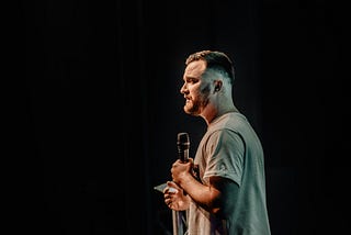 from the side, looking at a man who is standing on a stage, holding a microphone, with the spotlight on him