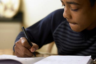 Black boy writing on paper with a pen. He’s wearing a blue shirt with stripes and appears to be thinking about what he’s writing.