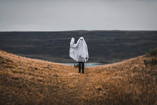 This image shows a person standing in a rural, grassy landscape dressed in a white ghost costume, reminiscent of a simple Halloween ghost with two eye holes cut out for visibility. They are on a path that looks to wind down a hill, with a body of water and hills in the distant background. The sky is overcast, contributing to a moody atmosphere.