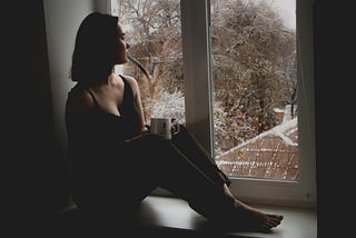 A woman holding a mug, sitting on a window seat, looking out into a snow covered landscape. She is silhouetted in darkness.