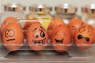 Eggs in an egg box. Each egg has a cartoon face drawn on it in black marker pen.