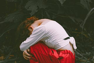A person wearing a white long-sleeve top and red pants crouches on the ground in a forest, head down and arms wrapped around their knees, surrounded by fallen leaves and dark green foliage, conveying a mood of sadness or introspection.