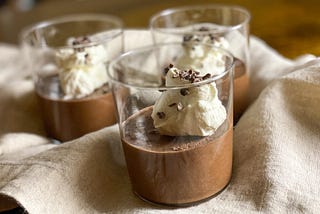 Three servings of chocolate mousse, served in glass tumblers, topped with whipped cream and cacao nibs