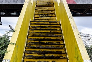 A photo of a steep vertical old, outdoor,  yellow staircase going up towards a bridge.