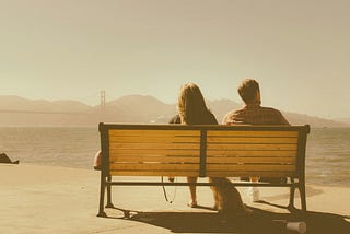 A couple sit on their bench and look at the water. Their dog sits between their legs.