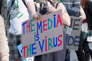 A protest crowd where five people can be seen carrying a variety of signs, with the most noticeable person holding a homemade sign that says “The Media is the Virus” in colorful letters.