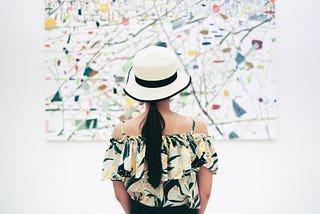 Image of a person with long hair in a ponytail wearing a colorful patterned top (blacks, yellows, some orange) of their shoulders. They are wearing a white hat brimmed with black. They are looking at a colorful splatter abstract painting that almost matches the top.