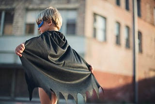 Little boy, back to the camera, holding out a black cape he is wearing. He also is wearing a blue mask.