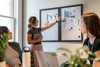 A woman holds a tablet and gestures toward a pinboard with design mockups and blueprints during a collaborative creative meeting, while two others look on.