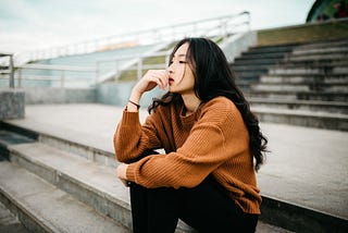 Photo of an Asian woman with long, black hair sitting on wide stairs and looking in the far distance ahead of her. She’s wearing black pants and a dark orange coloured long-sleeve sweater. Her left arm rests across her knees, and her right elbow rests on her right knee, her hand up near her mouth. The background is concrete stairs with some glass railings, maybe bleachers. It looks empty and bleak. No one else is around. It’s daylight, but cloudy or grey. She’s alone.