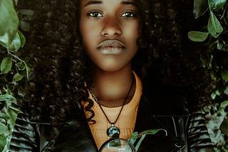 A close up of a black girl with full, curly hair and a black jacket staring into the camera. She is holding up a magnifying glass to her chest. There is green foliage around her.