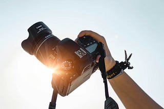 A hand holding an analogue camera in front of the sun with the sky in the background