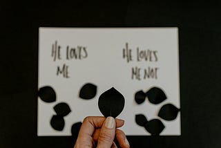 Picture of a white card with He Loves Me, He Loves Me Not written on it. Black petals on each side.