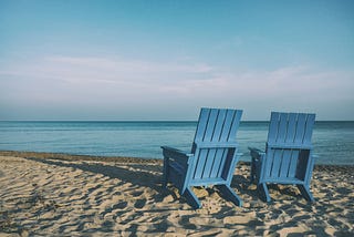 Two emtpy adironback chairs on the beach facing the ocean