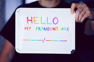 A woman holding a piece of white cardboard saying “HELLO, My PRONOUNS ARE — — — “ in rainbow colours