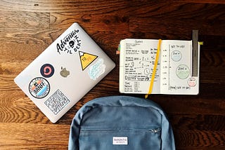 Mesa com mochila, computador e um caderno aberto.
