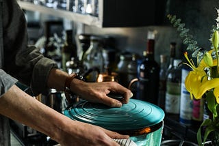 A man standing in his kitchen at the stove, under a range hood lifting an aqua coloured hot casserole pot off  with a tea towel.
