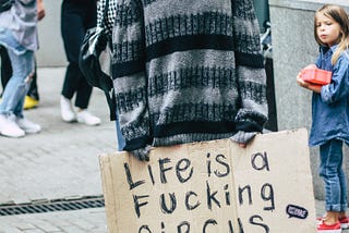 Man in clown makeup including a red-orange wig and a sweater. He has a blank expression on his face and a sign on cardboard that says “Life is a fucking circus.”