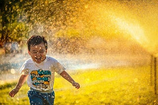A medium-skinned boy running gleefully through a sprinkler on the lawn. He has black hair swooping down towards the right and is wearing a white tee and blue and grey swim trunks. His shirt reads: “This world is full of hope. Let’s begin to proceed with peace.” The photo has a glowy light to it.