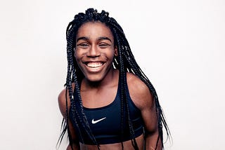 A teenage Black girl with long, thin braids laughs as she stands against a white background. She wears a black Nike cropped running tank.