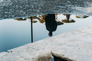 Reflexo em uma poça d’agua que espelha, de cabeça para baixo, a imagem de um homem andando na rua com uma máscara cobrindo a boca