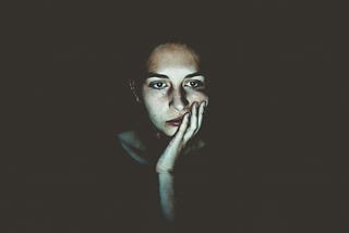 woman sitting in the dark, head resting in her hand, staring boredly at a screen.