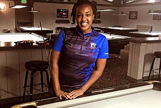 The author, Amani Ali, poses with her hands resting on the edge of a pool table with grey cloth in an empty pool room.