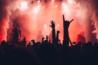 The image depicts the crowd at a concert. The stage is shrouded in smoke and red lights. You can see the silhouettes of the four band members. The audience takes up the lower third of the image frame. You have see hands making rocker signals and some people recording the artisit.