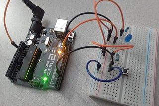 An Arduino board has wires connecting it to a breadboard. The breadboard has many wires, a button, and a light bulb.