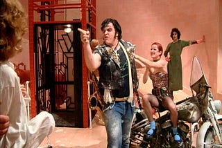 Zombie Eddie dismounts his motorcycle to point an angry finger at something off-camera, while his ex-girlfriend paws at him. Tim Curry in drag watches from the background.