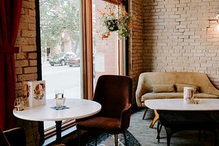 photo of inside of coffee shop, with two maroon velvet chairs around a small, round white table in the foreground and a beige couch and white coffee table in the background, with a large window on the left side of the picture with a plant with pink flowers hanging in the window.