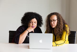 Two women sit at a deskand look at a laptop together.