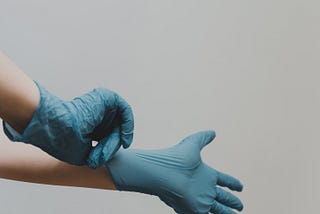 2 hands in blue surgical gloves. One gloved hand is pulling the other glove above the wrist.