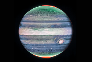 Image of Jupiter in white, blue, and green false color. Red patches at the north and south pole are also present. The planet is suspended on an empty black background.