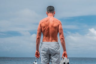 A man in a bathing suit stands looking out over the ocean, his bare back and shoulders facing the camera lens, with snorkeling gear grasped in his hands.