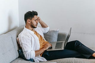 Man in a white shirt sits on a couch looking at his laptop.
